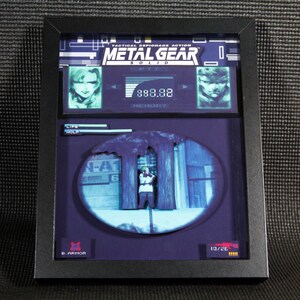 May include: A framed 3D diorama of a scene from the video game Metal Gear Solid. The diorama features a character standing in a forest, with a blue and green background. The diorama also includes a life meter, a memory meter, and a weapon selection screen. The text "Tactical Espionage Action Metal Gear Solid" is displayed at the top of the diorama.