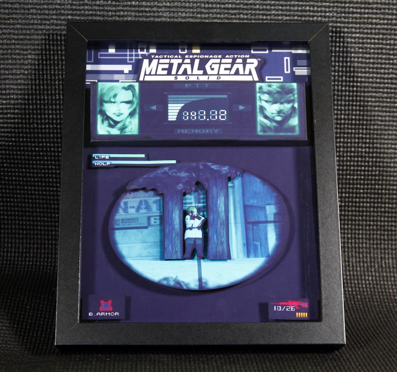 May include: A framed 3D diorama of a scene from the video game Metal Gear Solid. The diorama features a character standing in a forest, with a blue and green background. The diorama also includes a life meter, a memory meter, and a weapon selection screen. The text "Tactical Espionage Action Metal Gear Solid" is displayed at the top of the diorama.