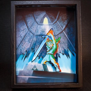 Op de afbeelding: Een ingelijst 3D-kunstwerk met een personage uit de videogame-serie The Legend of Zelda. Het personage, gekleed in groen en wit, houdt een zwaard omhoog in een blauw-grijze omgeving. Het kunstwerk is ingelijst in een zwart frame.