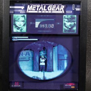 Op de afbeelding: Een ingelijst 3D-diorama van een scène uit de videogame Metal Gear Solid. Het diorama toont een personage dat een pistool vasthoudt en achter een boom staat. Het diorama is geplaatst tegen een blauwe en witte achtergrond. De lijst is zwart en heeft een glazen voorzijde. De titel "Metal Gear Solid" is op de lijst gedrukt.