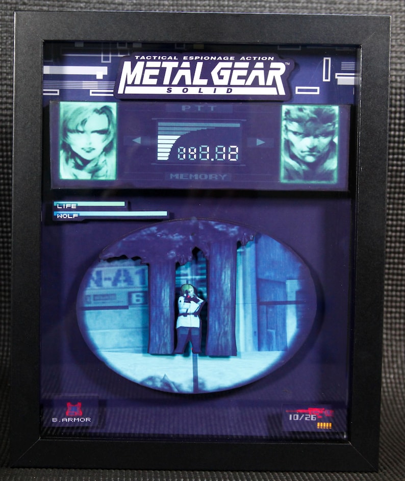 May include: A framed 3D diorama of a scene from the video game Metal Gear Solid. The diorama features a character holding a gun, standing behind a tree. The diorama is set against a blue and white background. The frame is black and has a glass front. The title "Metal Gear Solid" is printed on the frame.