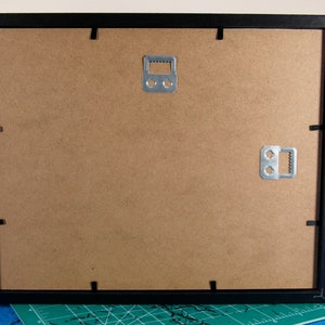 May include: Black picture frame with a brown cardboard backing. The frame has two metal hanging hooks on the back.