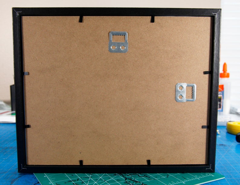 May include: Black picture frame with a brown cardboard backing. The frame has two metal hanging hooks on the back.
