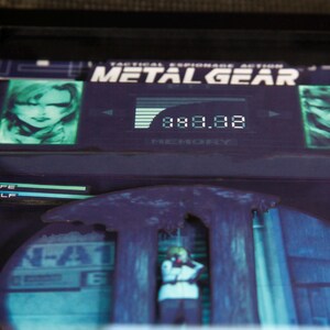 May include: A video game cartridge for the Metal Gear video game. The cartridge is black with a green screen showing a character's face and the game's title. The screen also displays the text "Tactical Espionage Action".