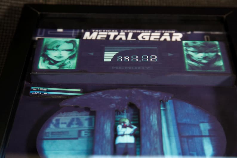 May include: A video game cartridge for the Metal Gear video game. The cartridge is black with a green screen showing a character's face and the game's title. The screen also displays the text "Tactical Espionage Action".