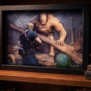 Puede incluir: Obra de arte enmarcada que representa una escena de un videojuego. La imagen muestra a un personaje enfrentándose a un gran monstruo con un tronco. Un reloj digital con el nombre "Leon" está en primer plano. La obra se exhibe en una estantería de madera.