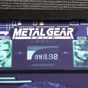 May include: A framed image of a video game screen from Metal Gear Solid. The screen shows the game's title, a character portrait, and a score of 888.88.