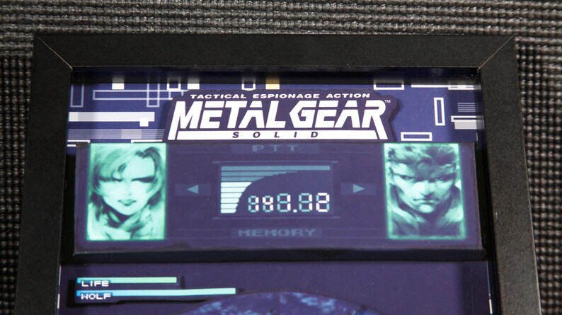 May include: A framed image of a video game screen from Metal Gear Solid. The screen shows the game's title, a character portrait, and a score of 888.88.
