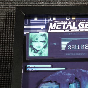 May include: A framed image of a video game screen from Metal Gear Solid. The screen shows a character's portrait, a health bar, and the text "Tactical Espionage Action Metal Gear Solid".