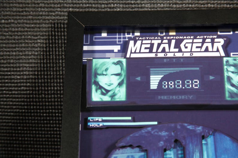 May include: A framed image of a video game screen from Metal Gear Solid. The screen shows a character's portrait, a health bar, and the text "Tactical Espionage Action Metal Gear Solid".
