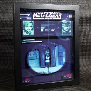 May include: A framed 3D diorama of a scene from the video game Metal Gear Solid. The diorama features a character standing in a hallway with the text "N-A1" and "6" on the wall. The diorama is set in a black frame with the game's title and logo at the top.