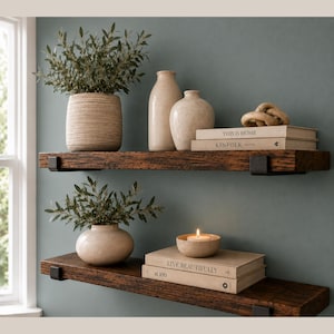 May include: Two dark brown wooden floating shelves mounted with black metal brackets. The top shelf holds a textured beige planter with greenery, two beige ceramic vases, and two books titled This is Home and Kinfolk. The bottom shelf holds a beige planter with greenery and a lit candle on two books.