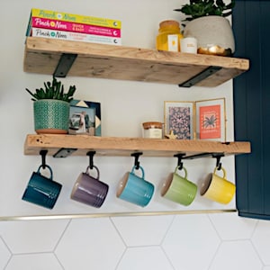 May include: Two rustic wooden shelves with black metal brackets are mounted on a white wall. The top shelf holds books, a potted plant, and a jar. The bottom shelf displays colorful mugs hanging below. The mugs are in shades of blue, purple, teal, green, and yellow.