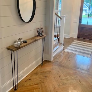 May include: A narrow, rustic wooden console table with black hairpin legs. The tabletop is a light brown color. Two glass jars with silver accents and a framed photo are on the table. A black oval mirror hangs on the wall above the table.