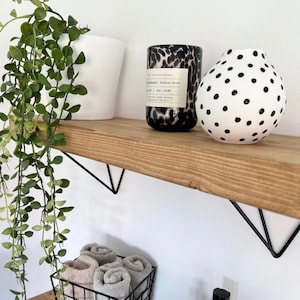 May include: A wooden shelf with a white wall background holds a black and white speckled candle, a white vase with black polka dots, and a white planter with a trailing green plant. Below, a wire basket contains rolled towels.