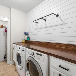 May include: A laundry room with white cabinets, a dark wood countertop, and a white shiplap wall. Two white washing machines are visible, along with a black pipe clothing rack. A small vase of flowers sits on the countertop.
