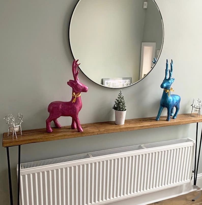 May include: A wooden console table with black hairpin legs, adorned with decorative deer figurines in pink and blue, each with a gold ribbon. A small potted Christmas tree and a round mirror complete the decor.