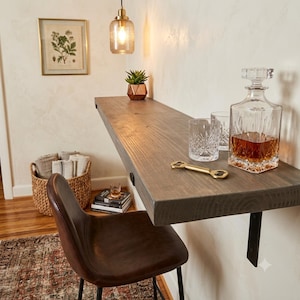 May include: A wall-mounted wooden bar shelf with a dark gray finish, supported by black metal brackets. On the shelf sits a clear glass decanter filled with amber liquid, two crystal whiskey glasses, and a gold-toned metal bottle opener.