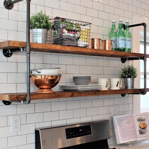 May include: Rustic, two-tiered wooden shelves supported by black metal pipes. The shelves are filled with kitchen items, including plants in metal pots, copper mugs, and plates. The shelves are mounted on a white tiled wall.