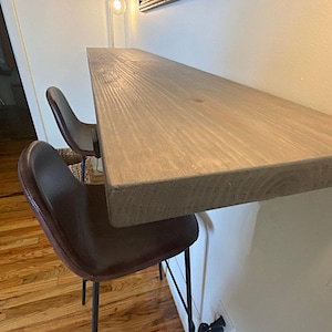 May include: A light brown wooden bar table is mounted to a white wall. Two brown leather chairs with black metal legs are positioned in front of the table. The table has a natural wood grain.