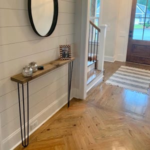 May include: A narrow, rustic wooden console table with black hairpin legs. The tabletop is made of light-colored wood. Two glass jars and a framed photo are displayed on the table. A black oval mirror hangs on the wall above the table.