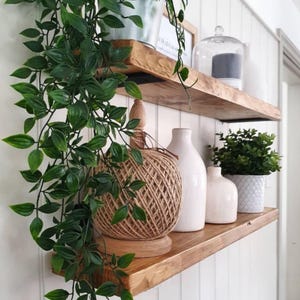 May include: Two wooden shelves with decorative items. A large ball of twine sits on a wooden base, next to white vases and a potted plant. Trailing green plants hang down from the top shelf.