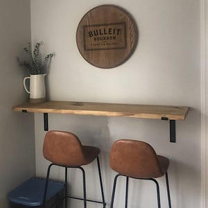 May include: A home bar setup featuring a wooden shelf with black metal brackets, two brown leather bar stools, and a Bulleit Bourbon sign. A white pitcher with greenery sits on the shelf. The sign is a round wooden piece with the brand name.