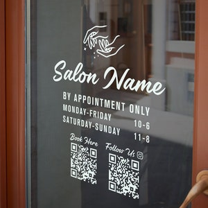 May include: White sign on a glass door with the text "Salon Name" and the opening hours. The sign also includes QR codes for booking appointments and following the salon on social media.