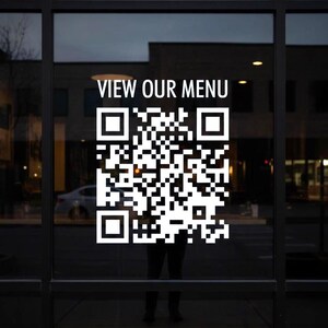 Może przedstawiać: Szklane okno z dużym białym kodem QR i napisem "VIEW OUR MENU". Kod QR to kwadratowy wzór czarno-białych kwadratów. Okno odbija otoczenie.
