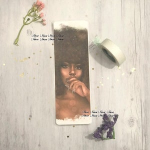 May include: A rectangular bookmark featuring a portrait of a woman with dark hair and skin, with the text "L'Amour" repeated along the edges. The woman is depicted with a thoughtful expression, her finger touching her lips. The bookmark is surrounded by decorative elements.