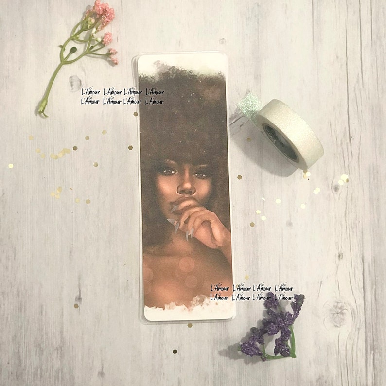 May include: A rectangular bookmark featuring a portrait of a woman with dark hair and skin, with the text "L'Amour" repeated along the edges. The woman is depicted with a thoughtful expression, her finger touching her lips. The bookmark is surrounded by decorative elements.