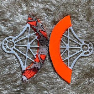 May include: Two white plastic structural frames with a webbed design, each featuring a curved fabric attachment. One attachment displays a bright orange and gray snakeskin pattern, while the other is a solid, vibrant neon orange fabric.