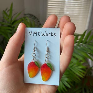 May include: A pair of mango-shaped earrings with red, orange, and yellow gradients, and a small green leaf. The earrings are on a light blue card with the text "MMCWorks". The earrings are suspended from silver-colored hooks.