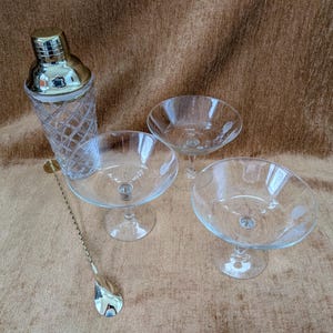 May include: A cocktail set featuring a silver-topped glass shaker with a diamond pattern, three clear coupe glasses, and a long bar spoon. The glasses have a wide, shallow bowl and a stem.