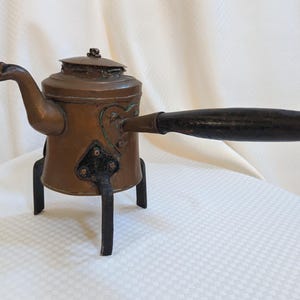 May include: Antique copper kettle with a dark wooden handle and a small lid. The kettle has a spout shaped like a bird's head and sits on a black metal stand with three legs. The kettle is approximately 8 inches tall.