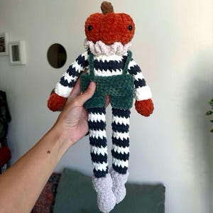 Könnte beinhalten: Handgemachte Plüschpuppe mit einem Kürbiskopf, die ein gestreiftes Hemd, grüne Latzhosen und gestreifte Beine trägt. Die Puppe hat einen Rüschenkragen und graue Füße. Die Puppe ist etwa 30 cm groß.