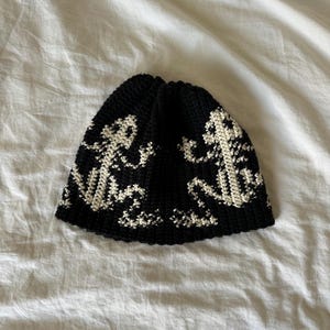 Può includere: Un berretto nero all'uncinetto con un motivo a forma di ancora color crema. Il cappello è realizzato con un filato morbido e strutturato e ha una corona arrotondata. Il motivo dell'ancora si ripete attorno alla base del cappello.