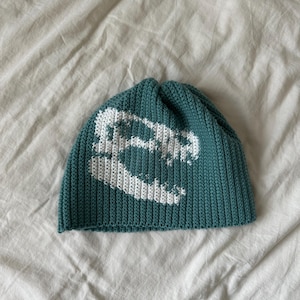 Peut inclure: Bonnet en tricot de couleur teal avec un motif de crâne de dinosaure blanc. Le chapeau a une texture côtelée et présente un motif détaillé d'un crâne de dinosaure sur le devant. Le bonnet est en matière douce et confortable.