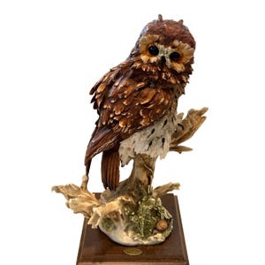May include: A detailed figurine of a brown and white owl perched on a textured tree branch. The owl has dark eyes and layered feather detailing. The branch is set on a square wooden base with a small plaque, featuring mossy green accents.