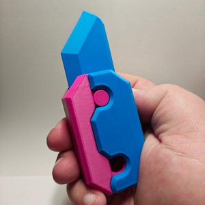 May include: A 3D printed utility knife with a bright blue blade and a handle featuring a combination of bright blue and magenta plastic components. The design includes a circular magenta accent piece and a cutout section on the lower handle area.