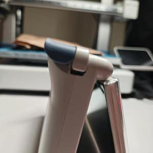 May include: A side profile of a light gray electronic device featuring a curved top edge with a dark blue accent piece. The device has a smooth, matte finish and a visible hinge mechanism connecting the upper section to a flat, dark screen panel.