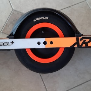 May include: A OneWheel+ XR electric skateboard with a black tire and orange accents. The board has a wooden deck and a black fender with the word "VEGA" printed on it. The board is designed for personal transportation.