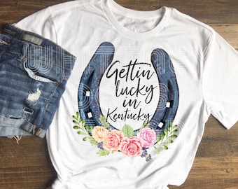 Może przedstawiać: Biała koszulka z niebieską podkową, kwiatowym wzorem i tekstem "Gettin' lucky in Kentucky".