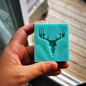 May include: A teal blue silicone mold with a deer head design. The mold is held in a hand.