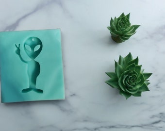 May include: A teal blue silicone mold with a cutout of an alien figure with one hand raised in a peace sign. Two small green succulent plants are on a white marble surface.