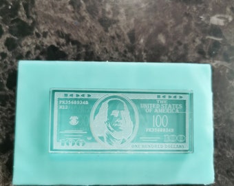 May include: A rectangular light blue silicone mold featuring a recessed design of a one hundred dollar bill. The impression shows the portrait of Benjamin Franklin, the text THE UNITED STATES OF AMERICA, 100, and the serial number FK3568934B.
