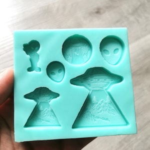 May include: A teal silicone mould with six different shapes: a UFO, a mountain range, an alien head, and three different alien figures.