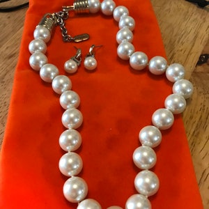 May include: A white pearl necklace and matching earrings set in a bright orange velvet pouch. The necklace is made of large, round pearls and has a silver clasp.