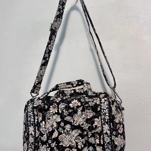 May include: Black floral print duffel bag with a detachable shoulder strap. The bag features a repeating pattern of flowers in shades of white, pink, and grey. It has a top handle and a side pocket. The bag is approximately 30 cm long.