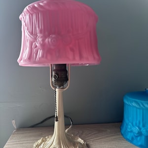 Puede incluir: Una lámpara de mesa vintage con una base blanca y una pantalla de vidrio rosa con un diseño floral. La pantalla tiene un borde festoneado y un remate decorativo.